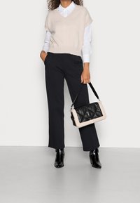 Lys beige strikket vest med v-udskæring, parret med skræddersyede sorte bukser og en sort quiltet taske med creme accenter, i en minimalistisk indstilling.