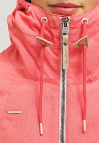 Gros plan d'une personne portant une veste à capuche rose corail avec une fermeture éclair frontale et des cordons, couvrant partiellement le bas du visage et les lèvres.