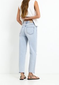 Ljusblå högmidjade jeans med en avslappnad passform, slitna nederkanter och bakfickor. Bärs med platta sandaler som har ljusbruna remmar.