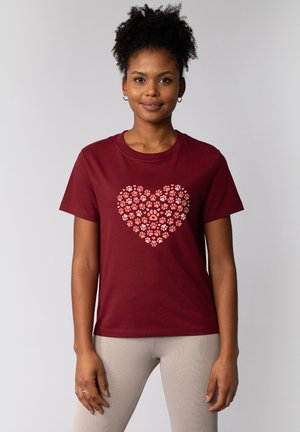 Femme aux cheveux bouclés portant un t-shirt bordeaux avec un cœur composé d'empreintes de pattes roses, associé à un pantalon côtelé beige clair.