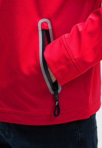 Mano parzialmente dentro di una tasca con zip di una giacca rossa brillante con rifiniture riflettenti e tirazip nero, indossata sopra pantaloni scuri.