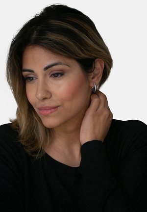 Gros plan sur une femme portant une petite boucle d'oreille en argent. Elle a de longs cheveux raides avec des mèches et se touche le cou.