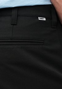 Μαύρο παντελόνι από υφασμάτινη υφή, με μπροστινή τσέπη και μια μικρή λευκή ετικέτα με το γράμμα "OBEY" κοντά στη μέση.