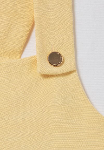 Tessuto giallo con una finitura текстurizzata, caratterizzato da dettagli con bottone dorato. Primo piano della zona della spallina che mette in risalto il colore e il materiale.