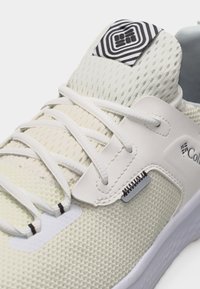 Κοντινή λήψη ενός λευκού αθλητικού παπουτσιού με πλέγμα ύφασμα, διασταυρωμένα κορδόνια και λογότυπο με ασπρόμαυρο σχέδιο στη γλώσσα.