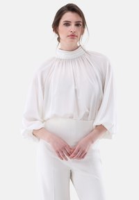 Blusa bianca, morbida e svasata con un colletto alto, maniche lunghe a palloncino e dettagli arricciati. Abbinata a pantaloni a vita alta in tono neutro.