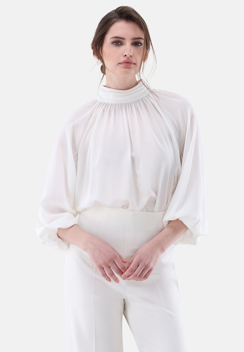 Blusa bianca, morbida e svasata con un colletto alto, maniche lunghe a palloncino e dettagli arricciati. Abbinata a pantaloni a vita alta in tono neutro.