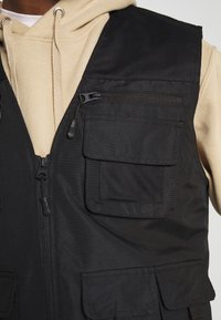 Sudadera beige con cordones llevada bajo un chaleco de utilidad negro que presenta múltiples bolsillos con cremallera y a solapa.
