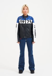 Giacca di pelle nera con accenti blu e bianchi, caratterizzata dai numeri "1971" in evidenza sul davanti. Abbinata a jeans a zampa d'elefante blu e stivali neri.