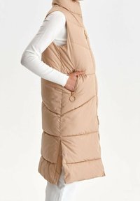 Wattierte Weste in hellem Beige mit hohem Kragen, Reißverschluss und Seitentaschen. Die Außenseite hat ein quilted Muster und eine glatte Textur.