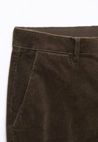 Plan rapproché sur un pantalon en velours côtelé marron foncé montrant la ceinture, la passants de ceinture et la poche latérale sur un fond blanc.