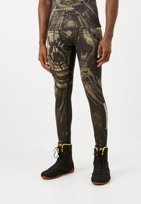 Male model wearing black and gold skull-patterned leggings and black high-top athletic shoes with red soles and yellow accents.