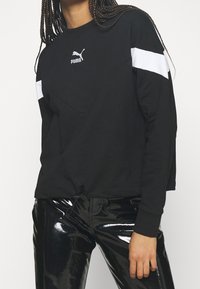 Zwarte sweatshirt met witte schouderaccenten en logo. Heeft een relaxte pasvorm en een textuurstof. Glanzende zwarte broek contrasteert met het bovenstuk.