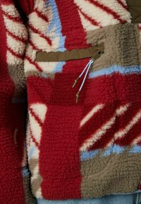 Veste en polaire rouge avec des motifs abstraits en crème, marron et bleu. Comprend une poche zippée et un cordon de serrage ajustable. Texture douce.