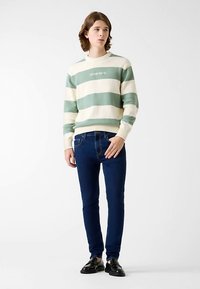 Pull en tricot rayé crème et vert, associé à un jean slim bleu foncé et des chaussures noires. Le pull est doté de poignets et d'un ourlet côtelés.