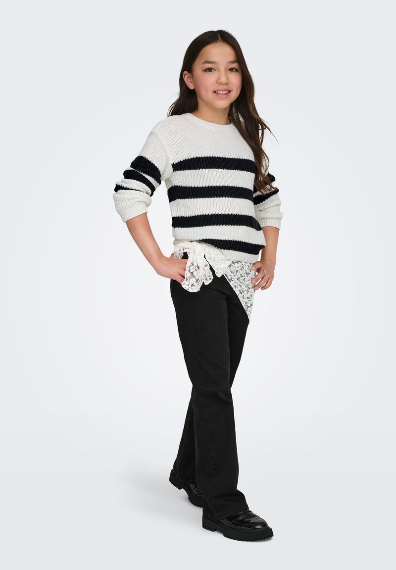 Fille debout, mains sur les hanches, portant un pull rayé blanc et noir, un pantalon noir, un détail de chemise en dentelle, et des chaussures noires sur un fond uni.