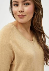 Maglione beige a scollo a V con una texture morbida, caratterizzato da dettagli cuciti lungo le spalle, indossato da un modello con lunghi capelli mossi.