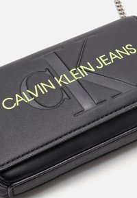 Borsa a tracolla in pelle nera con logo "CK" in rilievo. Presenta il testo audace "CALVIN KLEIN JEANS" in verde, una tracolla a catena e una superficie testurizzata.