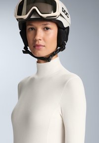 Bijela majica dugih rukava osnovnog sloja s visokim ovratnikom, glatke teksture i uskog kroja. Nošena s svijetlom skijaškom kacigom i naočalama.