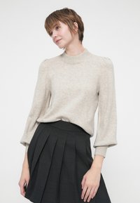 Sweater bege claro com gola alta, punhos canelados e mangas bufantes, combinado com uma saia pregueada escura. Textura suave, design minimalista.