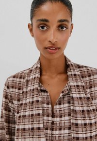 Bruine en witte geruite shirt met een zachte textuur, met knopen en een kraag, geopend gedragen over een bijpassend kledingstuk.