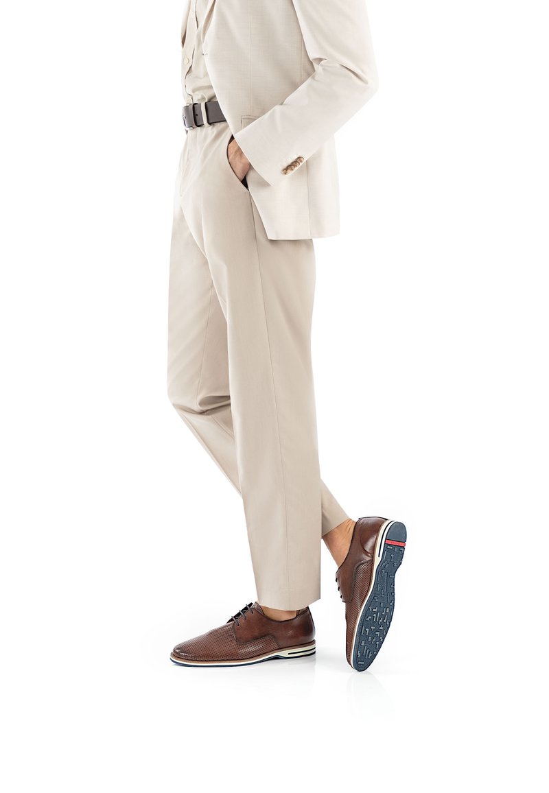 Braune Lederschuhe mit perforierten Details und weißem Akzentstreifen, kombiniert mit beigen Hosen und einem passenden Blazer. Klassischer Stil.