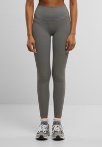 Leggings taille haute, moulants en matière grise mate, avec une texture extensible. Associés à des chaussures de sport grises et blanches.