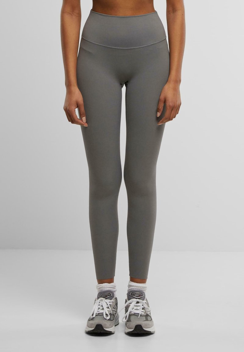 Leggings taille haute, moulants en matière grise mate, avec une texture extensible. Associés à des chaussures de sport grises et blanches.