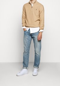 Beżowy sweter z zamkiem półtorągłym nałożony na białą koszulę, w połączeniu z jasnoniebieskimi dżinsami i białymi sneakersami. Sweter posiada małe logo.
