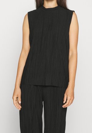 Femme portant un haut noir plissé sans manches associé à un pantalon noir plissé assorti, debout devant un fond clair uni.