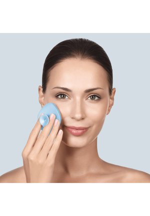 Kvinde med trukket hår bruger en blå silikone ansigtsrensningsbørste på sin højre kind mod en lys baggrund.