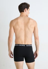 Männlicher Model steht mit dem Rücken zur Kamera und trägt schwarze Björn Borg Boxershorts vor einem schlichten weißen Hintergrund.