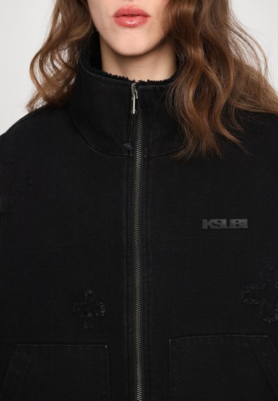 Black denim jacket with high collar, silver zipper, two front pockets, and embossed logo. Features textured patterns on the fabric.