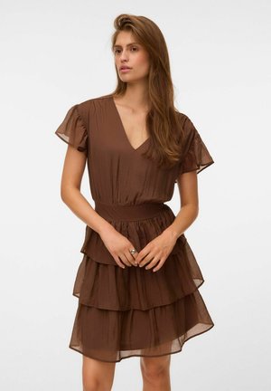Femme aux longs cheveux portant une robe marron à couches avec de courtes manches volantes, debout devant un fond uni.