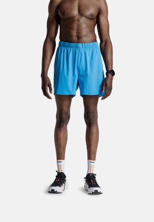 Shirtloze atletische man die felblauwe hardloopshorts draagt, witte sokken met oranje strepen, zwart-witte hardloopschoenen en een zwart polshorloge.