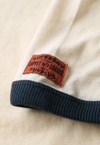 Detalle de camisa de algodón con un cuerpo crema, cuello de canalé en azul marino, y una etiqueta naranja con texto negro que dice "SUPERDRY SUPPLY GOODS."