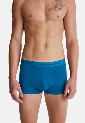 Torse masculin portant des boxers bleus avec un élastique portant le texte "United Colors of Benetton", avec un tatouage floral visible sur le côté droit de la poitrine.
