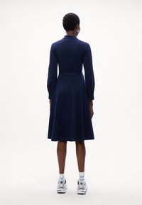 Abito blu navy con maniche lunghe, corpino attillato, gonna flare e cintura in vita; indossato con sneakers bianche e nere e calzini a fantasia.