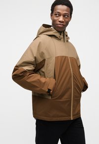 Καφέ και ταμπά μπουφάν με έντονα μπλοκ χρωμάτων· διαθέτει κουκούλα, φερμουάρ και μπροστινές τσέπες. Το υλικό φαίνεται ελαφρύ και με υφή.