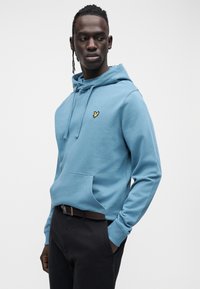 Sudadera con capucha azul claro de algodón con bolsillo tipo canguro, capucha con cordón y pequeño emblema amarillo en el pecho. Combinada con pantalones oscuros.