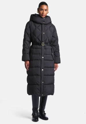 Femme portant un long manteau matelassé noir avec un col montant et une ceinture, associé à un pantalon noir et des bottines.