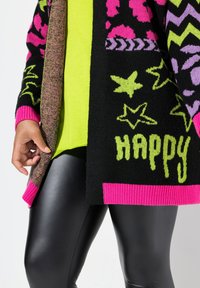 Cardigan nero con motivi rosa, verdi e viola, con la scritta "HAPPY". Indossato sopra una maglietta verde lime brillante e leggings neri lucidi.