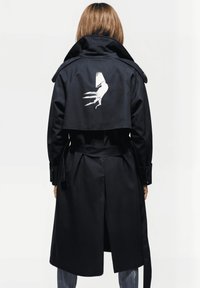 Persona con cabello hasta los hombros que lleva un abrigo negro, con un gráfico de una mano blanca en la parte superior de la espalda, de pie contra un fondo blanco.