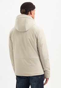 Beige gewatteerde jas met een hoge kraag en capuchon, met een gladde textuur. De achterkant toont een naaddetail en heeft een ontspannen pasvorm.