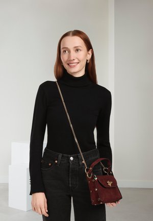 Burgunderrote Wildleder-Handtasche mit goldenen Akzenten, runden Beschlägen und einem oberen Griff. Modell trägt einen schwarzen Rollkragenpullover und hoch taillierte schwarze Jeans.