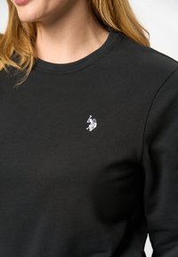 En kvinna bär en svart sweatshirt med en liten vit logotyp. Hon har axellångt hår och fokus ligger på hennes överkropp och halslinje.