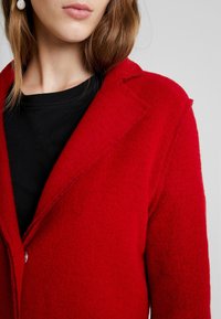 Rödullskappa med en texturerad yta, strukturerad krage och knappknäppning, parat med en svart skjorta under.