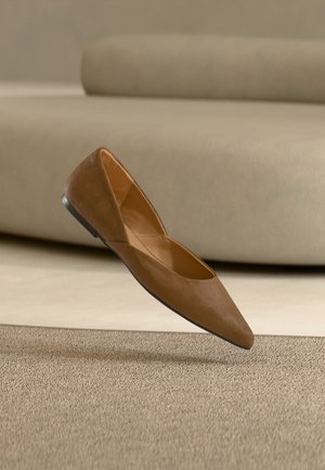 Chaussure plate en daim marron à bout pointu flottant au-dessus d'un tapis beige avec un canapé beige assorti flou en arrière-plan.