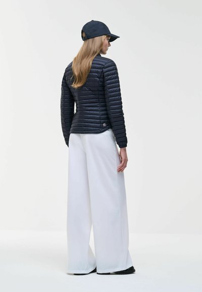 Woman wearing black quilted jacket, black cap, wide-leg white pants, and black shoes, standing facing sideways against white background.