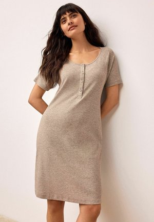 Femme aux longs cheveux noirs portant une robe beige côtelée à manches courtes avec des boutons, debout contre un mur de couleur claire.